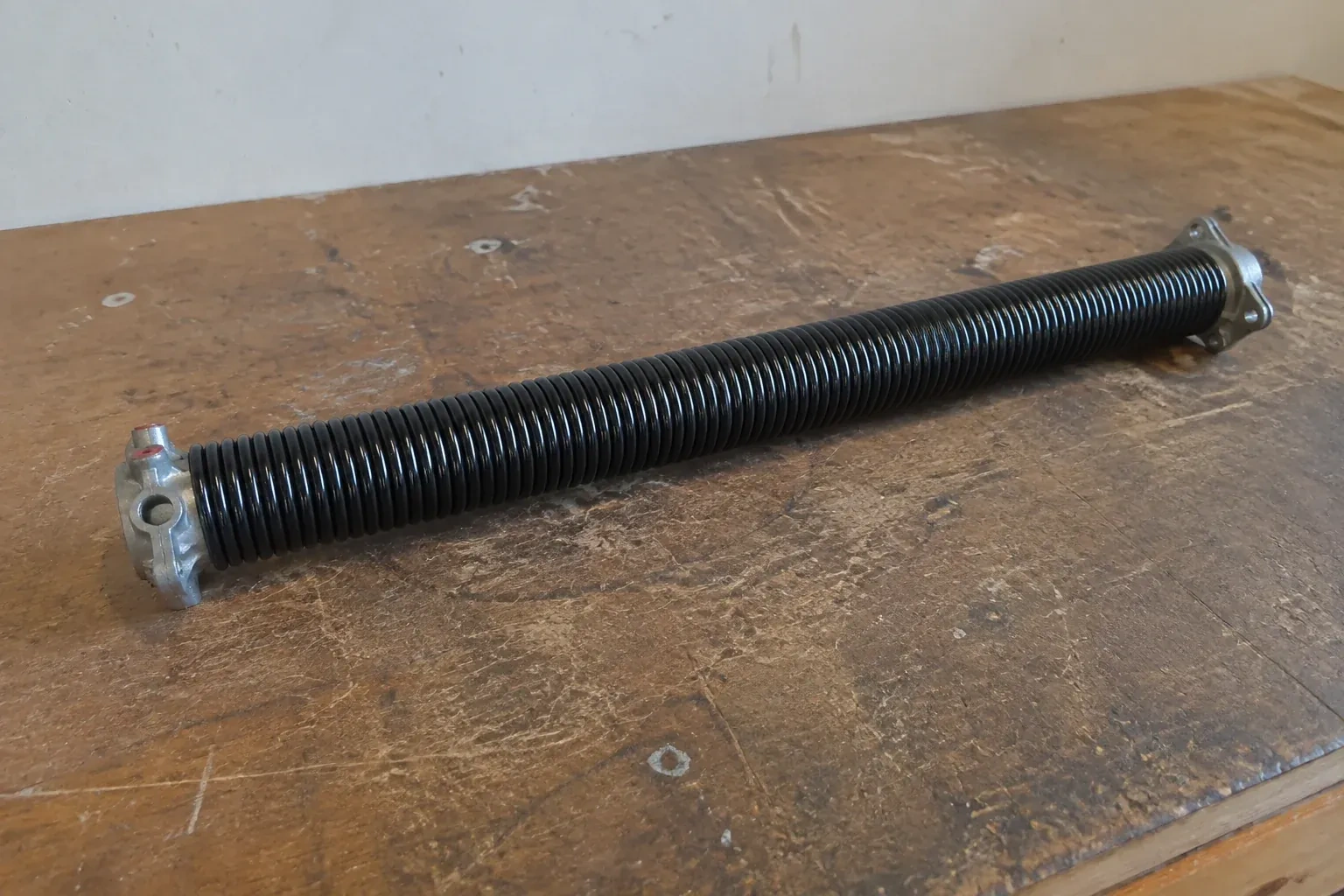 Detached black coil spring with metal end fittings on a workbench. — Torsion Spring Replacement, Vaughan