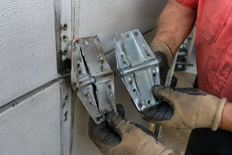 Gloved hands holding two metal garage-door hinges side by side, one discolored with a visible crack and the other bright silver. — Garage Door Hinge Replacement, Vaughan