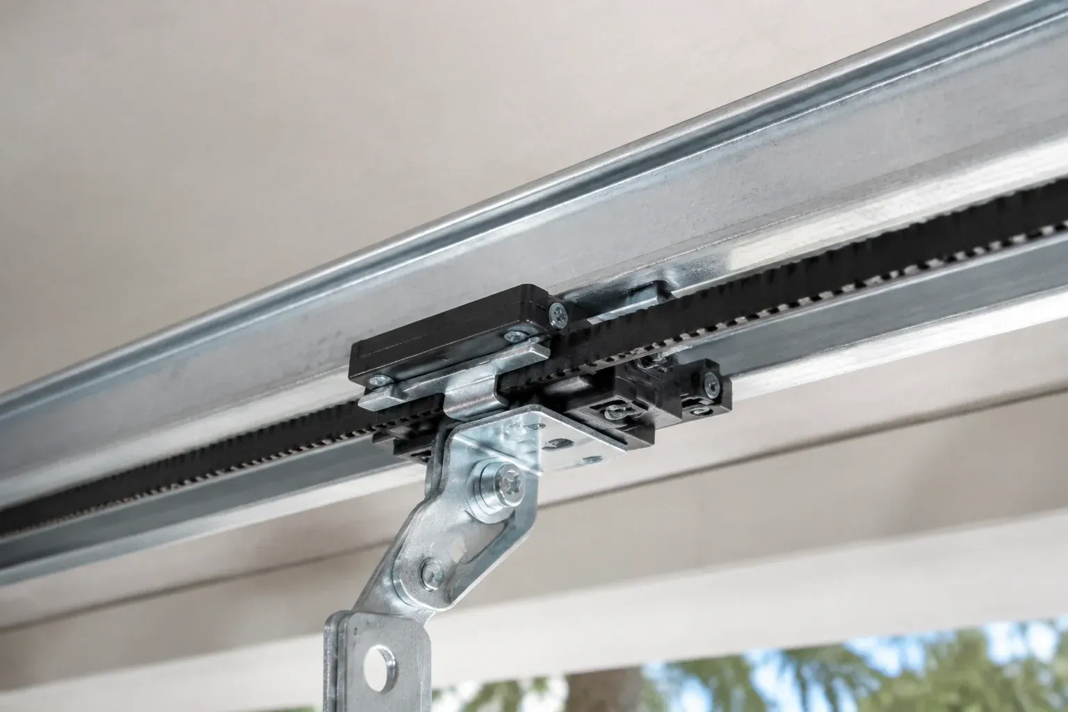 Black plastic trolley and bent galvanized bracket on a steel opener rail with a toothed black belt, against a ceiling. — Belt-Drive Opener Repair, Pine Grove