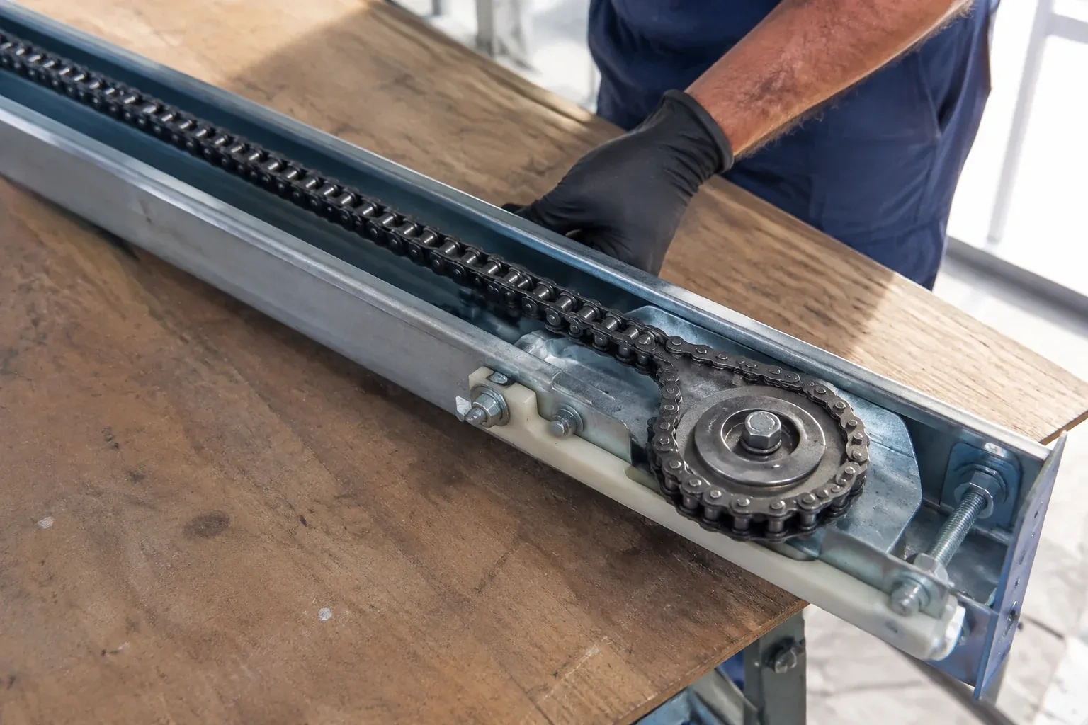 Gloved hand resting on a steel opener rail laid on a wooden bench, showing a drive chain looped around a sprocket wheel. — Chain-Drive Opener Repair, Kleinburg